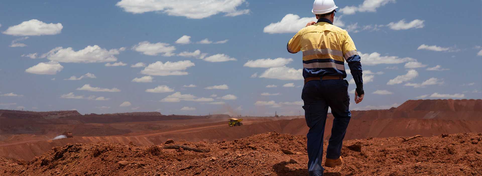 Worker in a hard hat and high-visibility shirt walking across red earth at an open-cut mine site, with a haul truck in the distance